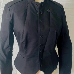 May include: A black jacket with a high collar and button-up front. The jacket has a tailored fit, with a fitted waist and decorative stitching details. The sleeves are long, and the jacket appears to be made of a smooth, woven fabric. The jacket is displayed on a mannequin.