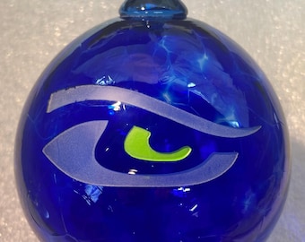 May include: A cobalt blue glass ornament with a stylized eye design in white and lime green. The ornament has a loop at the top for hanging. The glass has a textured appearance.
