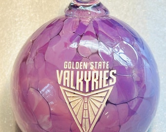 May include: A purple glass ornament with the text "GOLDEN STATE VALKYRIES" in white. The ornament has a textured surface and a clear glass swirl at the top.