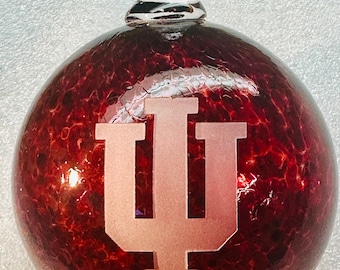 May include: A round, red glass ornament with a clear glass loop at the top. The ornament features a frosted, light-colored "IU" logo. The red glass has a speckled appearance.