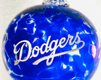 May include: A blue and white glass Christmas ornament with the word "Dodgers" in silver script. The ornament has a textured surface and a clear glass loop for hanging. A decorative item for sports fans.