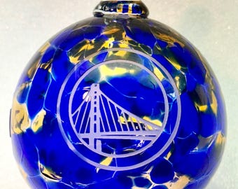 May include: A blue and gold glass Christmas ornament with a white etched design of the Golden State Warriors logo. The logo features a bridge and the team's name.