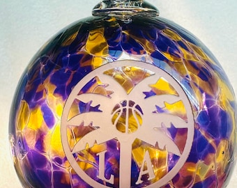 May include: A glass ornament with a clear loop and a round body. The ornament features a design with the letters "LA", a basketball, and palm trees. The background is a mix of purple, yellow, and gold.