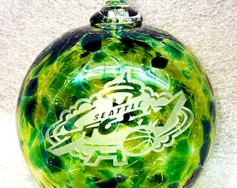 May include: Green glass Christmas ornament with a white Seattle SuperSonics logo etched on the front. The ornament has a green and black marbled design.