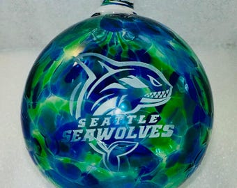 May include: A blue and green glass Christmas ornament with a white etched image of a shark and the text "Seattle Seawolves".