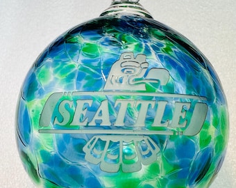 May include: A blue and green glass ornament with a white etched design of the Seattle skyline and a bird. The ornament has a clear glass loop for hanging.