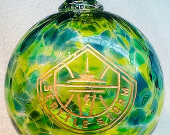 May include: A hand-blown glass ornament with a green and yellow mottled pattern. The ornament features a gold-colored emblem with the text "SEATTLE STORM" and a lightning bolt design. The ornament has a clear glass loop for hanging.