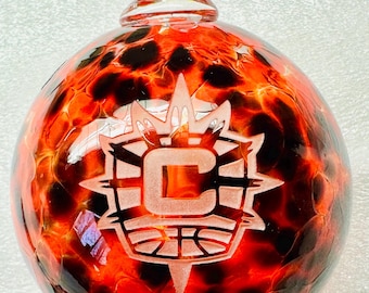 May include: A clear glass Christmas ornament with a fiery red and black pattern. The ornament features a team logo with the letter "C" in the center. The ornament has a clear glass loop for hanging.