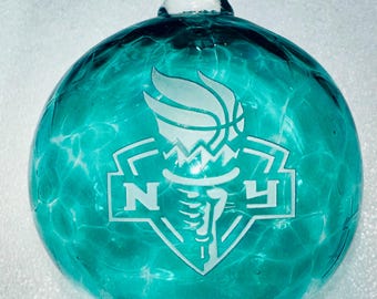 May include: A teal blue glass ornament with a white etched design of a basketball and the letters "NY" inside a shield.