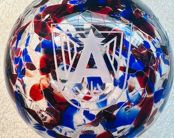 May include: A clear glass sphere with a mosaic pattern of red, blue, and white fragments. A white emblem with a star and the letter "A" is etched on the surface. The sphere is a decorative item.