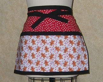 Pode incluir: Um avental vermelho com bolinhas brancas, um laço preto e uma estampa de homens de gengibre. O avental tem uma borda preta e uma cintura preta.