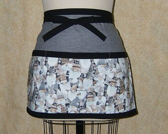 Pode incluir: Um avental cinza e preto até a metade com um laço preto para amarrar. O avental tem um bolso e uma borda inferior com uma estampa de gnomo.