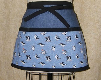 Pode incluir: Um avental pela metade com uma parte superior em jeans azul e uma parte inferior estampada com pinguins. A parte inferior apresenta um fundo azul claro com pinguins brancos e uma borda preta. O avental tem laços pretos.