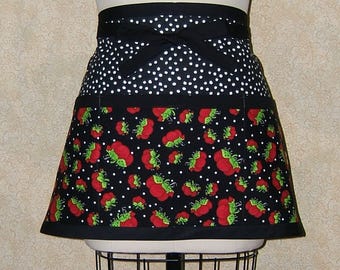 Pode incluir: Avençal de bolinhas brancas e pretas com uma cintura preta e uma borda inferior preta. O avental tem um bolso grande na frente com uma estampa de morangos.