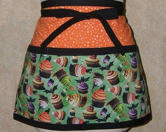 Pode incluir: Um avental de meia cintura com tema de Halloween com laço preto. A seção superior é laranja com bolinhas brancas, e a parte inferior apresenta um fundo verde com desenhos de cupcakes e morcegos. Acabamento preto.