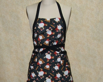 Pode incluir: Um avental preto com um padrão festivo de Papai Noel de desenho animado. O avental tem dois bolsos e alças pretas.