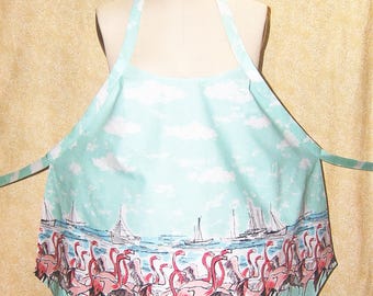 Pode incluir: Avental azul claro com um design de flamingos e veleiros. O avental apresenta uma cena de céu e água com nuvens brancas, flamingos cor-de-rosa e veleiros. O avental tem uma alça de pescoço e laços na cintura.