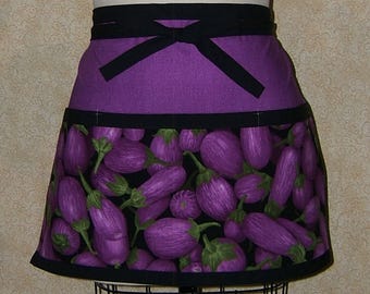 Pode incluir: Um avental roxo e preto com uma fita preta. O avental apresenta um padrão de beringelas roxas sobre um fundo preto. A seção superior é roxa sólida.