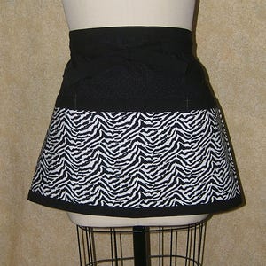 May include: A black and white zebra print apron with a black waistband and trim. The apron is displayed on a mannequin. The zebra print is on the lower half of the apron.