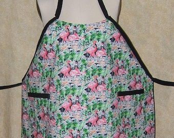 Pode incluir: Um avental com um padrão de flamingo. O avental apresenta um padrão repetido de flamingos rosa num cenário tropical com folhagem verde. Possui acabamentos pretos, uma alça de pescoço preta e dois bolsos pretos.