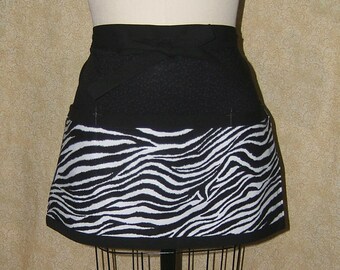 Pode incluir: Um avental com estampa de zebra em preto e branco. O avental tem uma cintura preta e uma bainha preta. A estampa de zebra está na metade inferior do avental. O avental é exibido em um manequim.