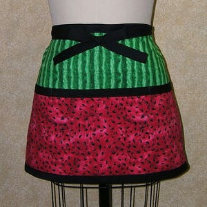May include: A black and green watermelon-themed apron with a black bow tie. The apron has a green and white striped top section and a red and black watermelon seed pattern bottom section.