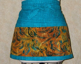 Pode incluir: Um avental curto azul-petróleo e castanho com um padrão de pássaros. A faixa superior e as amarrações são azul-petróleo, enquanto a seção inferior apresenta um desenho repetido de pássaros em tons de castanho, azul-petróleo e amarelo.