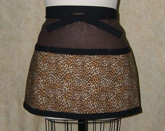 Pode incluir: Um avental marrom e com estampa de leopardo com cós e acabamento pretos. O avental é amarrado na cintura com um laço preto. O padrão de estampa de leopardo é uma mistura de manchas bege, marrons e pretas.