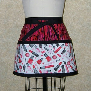 May include: A black and pink apron with a white bottom panel featuring a pattern of lipstick, nail polish, and makeup brushes.