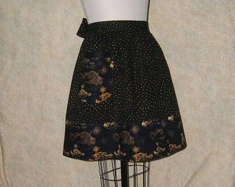 Pode incluir: Um avental preto com bolinhas douradas e um bolso com uma estampa de peixe-pescador em preto e dourado. O avental tem uma borda preta e dourada.