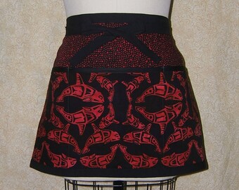 Pode incluir: Avental preto com um design de orca vermelha. O avental apresenta uma faixa de cintura e laços pretos, com um padrão de pontos vermelhos na seção superior. A seção inferior exibe um padrão repetido de orcas estilizadas.