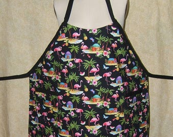 Pode incluir: Avental preto com estampa tropical de flamingos. O avental apresenta um padrão repetido de flamingos rosa, palmeiras, cabanas e flores. Possui uma alça de pescoço preta e laços na cintura, e é exibido em um manequim.