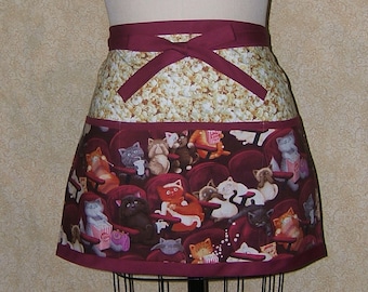 Pode incluir: Avental bordô com estampa de pipoca e gatos no cinema. A parte superior apresenta um padrão de pipoca, enquanto a inferior mostra gatos em cadeiras de cinema. Ideal para amantes de gatos e culinária.