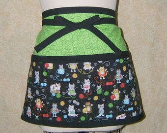 Pode incluir: Um avental curto com laço e acabamento pretos. A seção superior é verde limão com um padrão de redemoinho. A seção inferior é preta com um padrão de gatos, novelos de lã e pegadas.