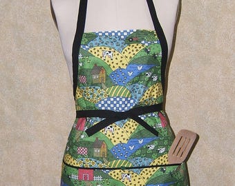 Pode incluir: Um avental colorido com uma estampa de cena de fazenda, apresentando casas, vacas e colinas em verde, amarelo e azul. O avental tem alças pretas e uma gravata preta na cintura, com uma espátula de madeira no bolso.