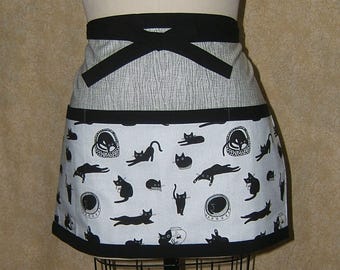 Pode incluir: Um avental com tema de gato em preto e branco. O avental apresenta um laço preto na cintura e uma borda preta. A parte inferior exibe um fundo branco com ilustrações de gatos pretos. A parte superior tem um padrão de listras cinza e branco.