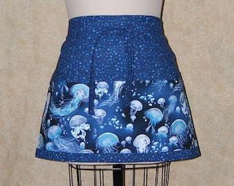 Pode incluir: Um avental azul com um design de água-viva. O avental apresenta um fundo azul escuro com bolinhas brancas na metade superior e uma borda de água-viva em tons de azul e branco. O avental é exibido em um manequim.