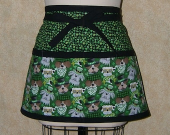 Pode incluir: Um avental verde com cós e laços pretos. A seção superior apresenta um padrão de pequenos trevos verdes sobre um fundo preto. A seção inferior exibe um padrão de cães usando chapéus e óculos de sol em um fundo verde.