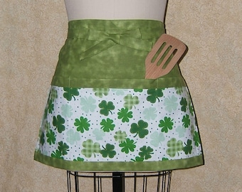 Pode incluir: Avental verde e branco com estampa de trevo e um bolso para uma espátula. O avental tem uma parte superior verde com uma estampa de trevo branco na parte inferior.