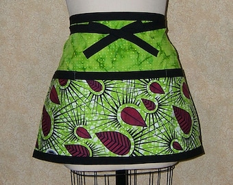 Pode incluir: Um avental verde e preto com um padrão de folhas vermelhas e brancas. O avental tem um laço preto para amarrar e um bolso.