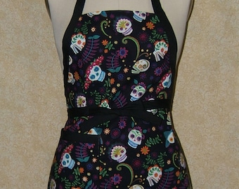 Pode incluir: Avental preto com um padrão colorido de caveiras de açúcar com flores e folhas. O avental tem uma alça de pescoço preta e laços na cintura. O design inclui detalhes brancos, azuis, verdes, roxos e laranja.