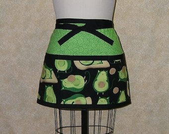 Pode incluir: Avental preto e verde com um padrão de redemoinho verde na parte superior e uma estampa de abacate preto e branco na parte inferior. O avental tem uma gravata preta.