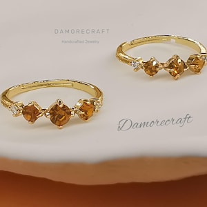 Puede incluir: Dos anillos de oro con tres piedras preciosas de color ámbar y pequeñas piedras transparentes. Los anillos están sobre una superficie blanca. El texto "Damorecraft Handmade Jewelry" es visible sobre los anillos.