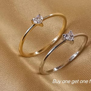 May include: Two thin rings, one gold-toned and one silver-toned, each featuring a small, clear, heart-shaped stone set at the top. The text "Buy one get one free" is printed in white in the lower right corner of the image.