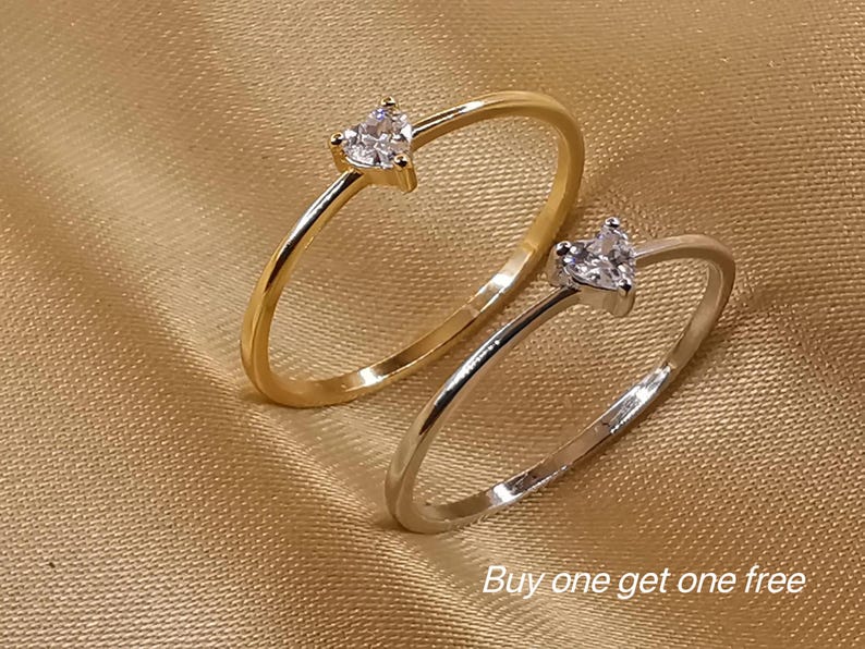 May include: Two thin rings, one gold-toned and one silver-toned, each featuring a small, clear, heart-shaped stone set at the top. The text "Buy one get one free" is printed in white in the lower right corner of the image.