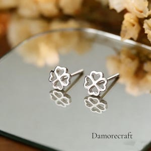 May include: Two silver stud earrings, each in the shape of a four-leaf clover. The earrings are presented on a reflective surface, with the brand name "Damorecraft" visible at the bottom.
