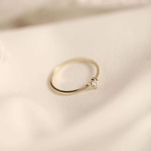 May include: A thin gold band ring featuring a single small, clear, faceted gemstone set in a prong setting. The ring has a polished metallic finish and a simple, circular design, showcasing a delicate and refined appearance in its construction.