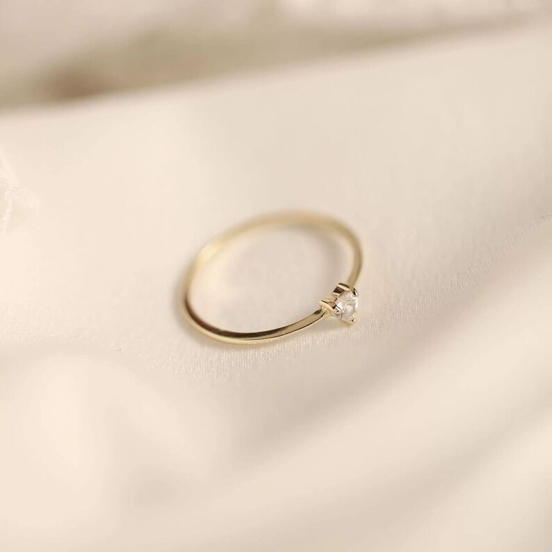 May include: A thin gold band ring featuring a single small, clear, faceted gemstone set in a prong setting. The ring has a polished metallic finish and a simple, circular design, showcasing a delicate and refined appearance in its construction.