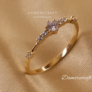 May include: A gold ring with a single, clear, round gemstone and smaller clear gemstones set along the band. The ring is on a beige fabric background.