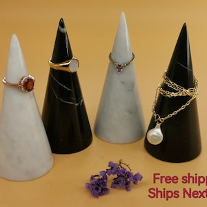 Puede incluir: Cuatro soportes cónicos para anillos y collares hechos de mármol blanco y negro. Dos soportes son blancos y dos son negros. Los soportes están dispuestos en una fila sobre una superficie beige.